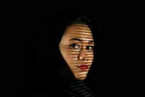 Young beautiful modern Malay woman headscarf black background with gobo cast shadow on face photo