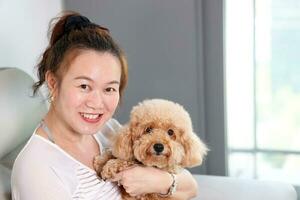 Beautiful attractive middle age southeast Asian woman sitting on sofa posing happy with small pet brown dog photo