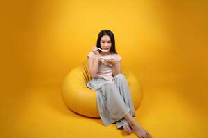 Beautiful young south east Asian woman holding empty chopstick soup spoon bowl pretend acting posing see eat taste smell feed offer satisfaction yummy siting on beanbag seat yellow orange background photo