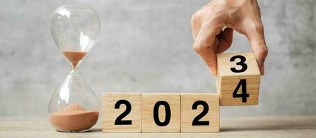 hand flipping block 2023 to 2024 text with hourglass on table. Resolution, time, plan, goal, motivation, reboot, countdown  and New Year holiday concepts photo