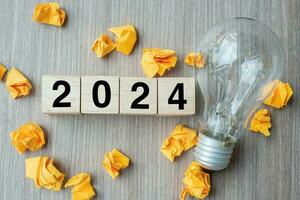 2024 texto madera cubo bloques y se desmoronó papel con bombilla en de madera mesa antecedentes. nuevo año nuevo ideas, creativo, innovación, imaginación, inspiración, resolución, estrategia y objetivo concepto foto