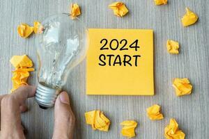 2024 Start words on yellow note and crumbled paper with Businessman holding lightbulb on wooden table background. New Year New Idea Creative, Innovation, Imagination, Resolution and Goal concept photo