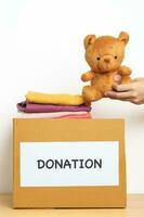 Kid Donation, Charity, Volunteer, Giving and Delivery Concept. Hand holding Bear doll and Clothes into Donation box at home for support and help poor, refugee and homeless people. Copy space for text photo