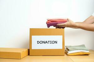 Donation, Charity, Volunteer, Giving and Delivery Concept. Hand holding Clothes into Donation box at home or office for support and help poor, refugee and homeless people. Copy space for text photo