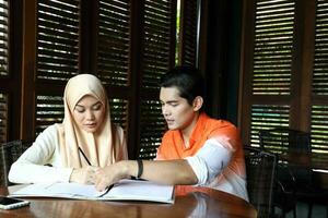 Young asian malay man woman with headscarf sit at rustic cafe table talk discuss business study read photo