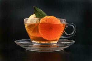 Liquid tea lemon orange slice green leaf cinnamon stick in transparent glass teacup saucer on black background photo