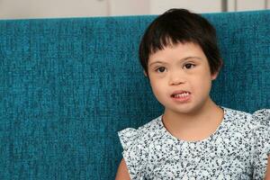 Sureste asiático pequeño pequeño niña niño sentado en azul sofá Mira contento sonrisa pose. ella tener abajo síndrome foto