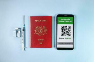 Digital international vaccine passport smart phone screen qr code app coronavirus covid 19 immunization red Malaysian passport small bottle disposable medical syringe needle on light blue background photo