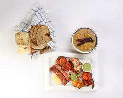 Mixed grill kebab chicken mutton beef prawn fish mint sauce lemon pickled onion bread basket chapati naan tandoori paratha dal lentil fry tadka copper pot on white dish over white background photo