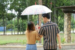 Young Asian Malay man woman couple outdoor green park walk talk discuss mingle under umbrella from behind photo