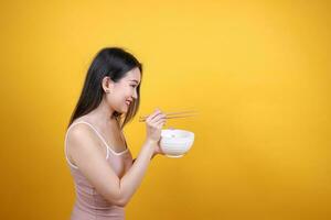 Beautiful young south east Asian woman holding chines empty chopstick Chinese soup spoon bowl utensil pretend acting posing see eat taste smell feed offer satisfaction yummy yellow orange background photo