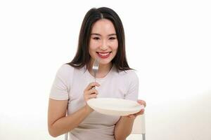 hermosa joven sur este asiático mujer fingir interino posando participación vacío tenedor cuchara blanco plato en mano comer gusto Mira ver blanco antecedentes sonrisa contento comida en boca foto