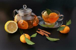 Liquid tea lemon orange slice green leaf cinnamon stick in transparent glass teacup saucer teapot kettle on black background photo