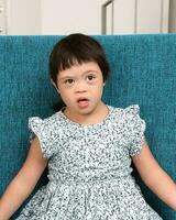 Sureste asiático pequeño pequeño niña niño sentado en azul sofá Mira contento sonrisa pose. ella tener abajo síndrome foto