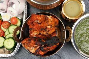 Spicy red Chicken grilled tikka tandoori nan bread green spinach curry yogurt sauce dal tomato cucumber onion salad set in metal stainless steel brass copper pot on black char wood background photo