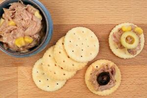 Tuna fish spread topping cracker biscuit tapas corn green black olive small bowl on wooden board over white background photo