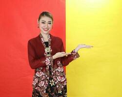 Asian woman traditional maroon kebaya sharong on red yellow paper background posing show display palm of hand photo