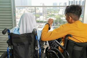 retrato de joven hombre mujer sur este asiático malayo sentado participación manos en silla de ruedas Mira fuera de terminado balcón ventana. paralizado cintura abajo. foto