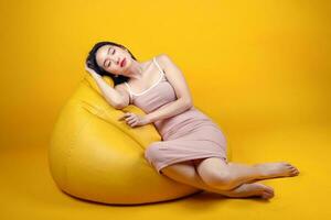 Beautiful young south east Asian woman sit on a yellow orange beanbag seat color background relax rest think emotion imagine expression pose sleep photo
