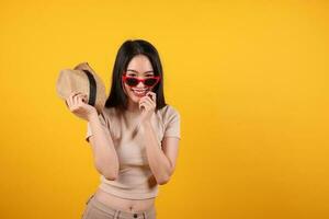 Beautiful young south east Asian woman red frame sunglass hat pose style fashion peak happy on yellow orange background holding hat in hand smile happy photo