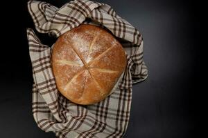 Fresh baked round bread loaf wrapped in checkered kitchen fabric napkin towel over black background photo