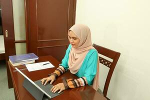 joven asiático malayo musulmán mujer vistiendo Pañuelo a hogar oficina estudiante sentar a mesa computadora libro archivo papel estudiar trabajo foto