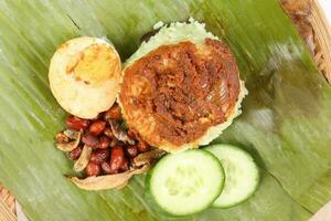 Nasi Lemak fragrant rice cooked in coconut milk served with sambal friend peanut anchovy boiled egg packed in banana leaf on round bamboo plate on white background photo