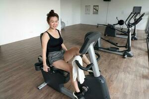 hermosa atractivo medio años Sureste asiático mujer hacer ejercicio estacionario bicicleta ciclo ejercicio bicicleta en un gimnasio sano estilo de vida Mira adelante a cámara foto