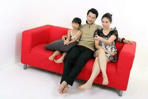 South East Asian young couple father mother daughter parent girl child on red sofa watch see use remote on white background photo