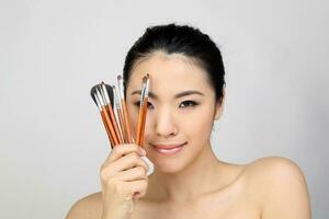 Young beautiful Southeast Asian woman beauty fashion makeup light grey white background holding makeup brush over eye face smile photo