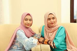 Two young Asian Malay Muslim woman wearing headscarf at home office student sitting on sofa talk mingle look at side other smile happy photo