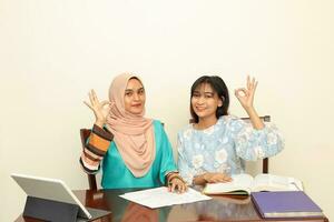 Two young Asian Malay Muslim woman wearing headscarf at home office student sitting at table talk mingle computer book document study ok finger sign look at camera photo