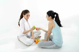 Two beautiful young south east Asian Chinese healthy workout exercise sit on floor holding fruits green apple orange banana talk disuses on white background photo