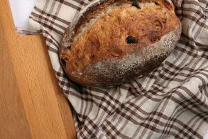 Rustic whole grain artisan bread loaf with cranberry raisin dry fruit nuts wrapped in checkers cloth with wooden chopping board over table top flat lay view photo