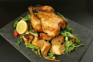 Whole roasted grilled chicken poultry bird with baked potato vegetable salad tomato lemon on black slate stone cutting board black background photo
