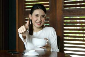 joven asiático mujer sentar a rústico café participación sopa fideos cuenco mano dedo pulgares arriba bueno firmar Mira frente a cámara atención en dedo foto