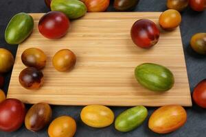 Red orange yellow green tomato mix variety on around wooden chopping board frame copy text space over black slate stone background photo
