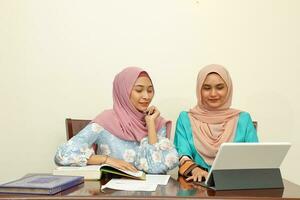 Two young Asian Malay Muslim woman wearing headscarf at home office student sitting at table talk mingle look at computer book document study discuss smile happy photo