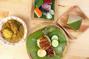 Nasi Lemak fragrant rice coconut milk with sambal friend peanut anchovy egg packed banana leaf round bamboo plate colorful nyonya kuih sweet desert palm leaf plate on wooden background photo