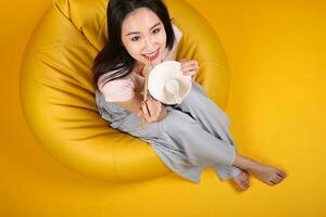 Beautiful young south east Asian woman holding empty chopstick soup spoon bowl posing see eat taste smell feed offer satisfaction yummy siting on beanbag seat yellow orange background top view photo