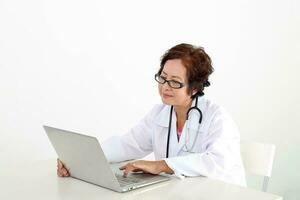 Elderly asian Chinese female doctor on white background wearing white apron stethoscope look typing working at laptop notebook pc photo