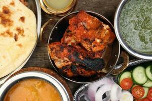 Spicy red Chicken grilled tikka tandoori nan bread green spinach curry yogurt sauce dal tomato cucumber onion salad set in metal stainless steel brass copper pot on rustic vintage wood background photo