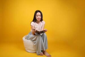 Beautiful young south east Asian woman sits on a white beanbag seat orange yellow color background pose fashion style elegant beauty mood expression relax smile look read book photo