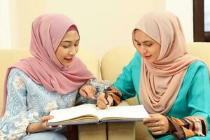 Two young Asian Malay Muslim woman wearing headscarf at home office student sitting on sofa talk mingle look at book document study discuss other smile happy photo