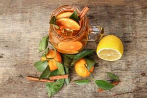 Liquid ice lemon orange tea with slice green leaf cinnamon stick in transparent glass jar mug on rustic wood background whole fruit photo