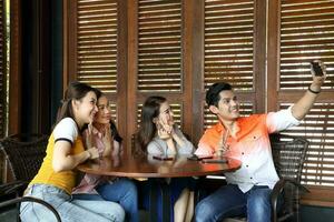 grupo joven asiático malayo hombre mujer a rústico de madera café mesa reunirse hablar discutir negocio estudiar selfie yo retrato mano inteligente teléfono foto
