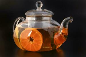 Liquid tea lemon orange slice green leaf cinnamon stick in transparent glass teapot kettle on black background photo