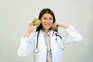 joven asiático hembra médico vistiendo delantal uniforme sayo estetoscopio participación señalando demostración comiendo sano verde manzana foto