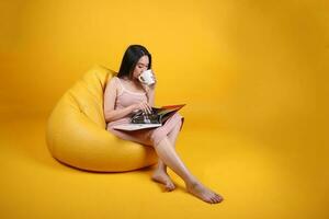 hermosa joven sur este asiático mujer sentar en un amarillo naranja bolsa de frijoles asiento color antecedentes relajarse leer estudiar pensar libro revista sostener blanco café taza foto