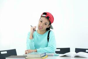 Yong Asian youth girl in casual dress cap sit at table book notebook pen look at camera on white background photo